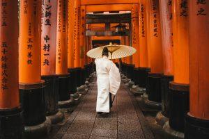 Personne en tenue traditionnelle marchant sous une allée de torii orange, perspective profonde et lumière douce. Person in traditional clothing walking under a corridor of orange torii gates with deep perspective.