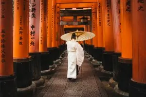 Personne en tenue traditionnelle marchant sous une allée de torii orange, perspective profonde et lumière douce. Person in traditional clothing walking under a corridor of orange torii gates with deep perspective.