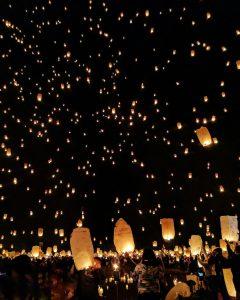 Milliers de lanternes lumineuses dans un ciel noir au-dessus d’une foule, scène de fête nocturne. Thousands of glowing lanterns in a dark sky above a crowd during a night festival.