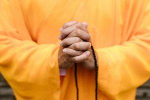 Mains jointes tenant un mala noir, robe orange en arrière-plan, geste de prière. Hands clasped holding a black mala, orange robe in the background, prayer gesture.
