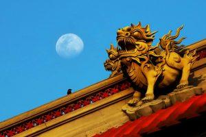 Sculptures de gardiens sur un toit de temple sous un ciel bleu, lune visible en arrière-plan. Temple roof guardian sculptures against a blue sky with the moon in the background.