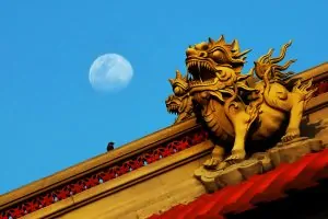 Sculptures de gardiens sur un toit de temple sous un ciel bleu, lune visible en arrière-plan. Temple roof guardian sculptures against a blue sky with the moon in the background.