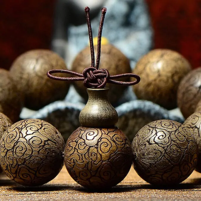 Close-up of authentic carved wooden mala beads bracelet with intricate golden cloud patterns, red silk cord, and traditional Tibetan vase charm, set against a serene Buddha statue background for spiritual protection and meditation – bracelet mala bouddhiste en bois sculpté, collier de prière tibétain artisanal.
