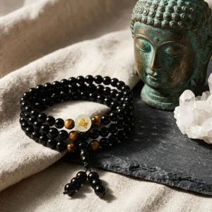 Collier mala 108 perles en obsidienne noire et oeil de tigre, medaillon zodiacal grave a l or, presente en still life avec une tete de bouddha en bronze et un cluster de quartz sur une ardoise